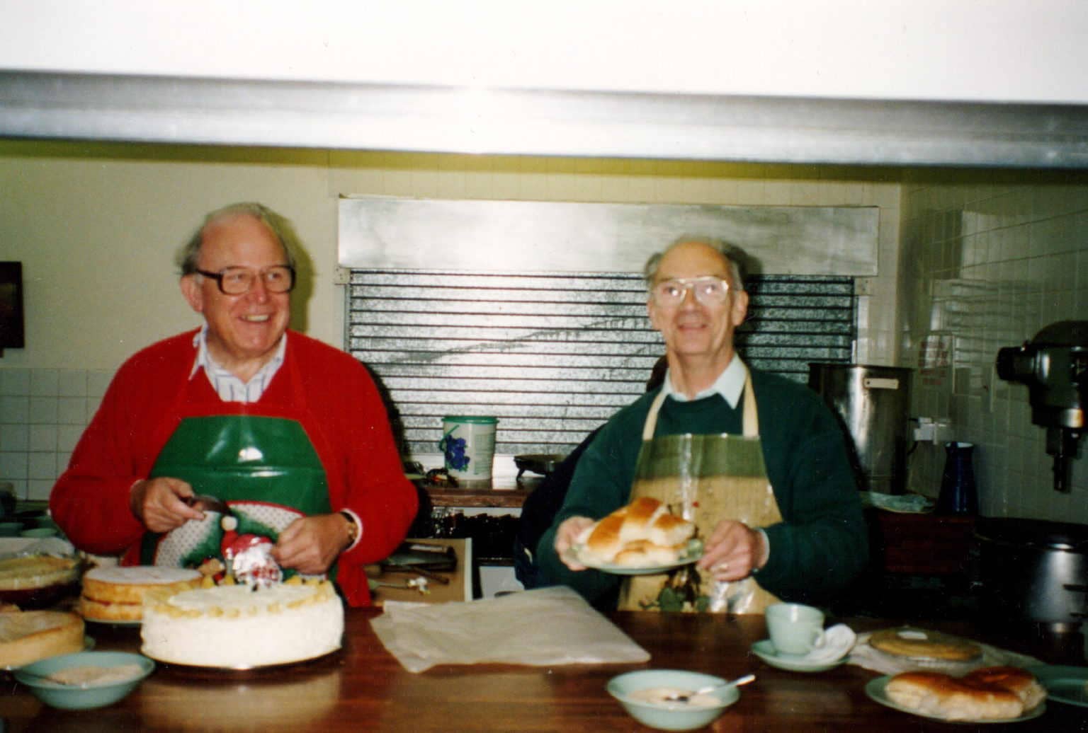 colour photo of two middle aged white men wearing glasses and smiling. They are in a kitchen with cakes.