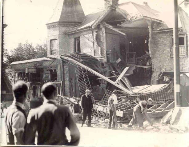 Black and white photo of a bombed building with the sign Pinecliff Pharmacy. The backs of two men are in the foreground, and there are three men starting to clear debris near the building.