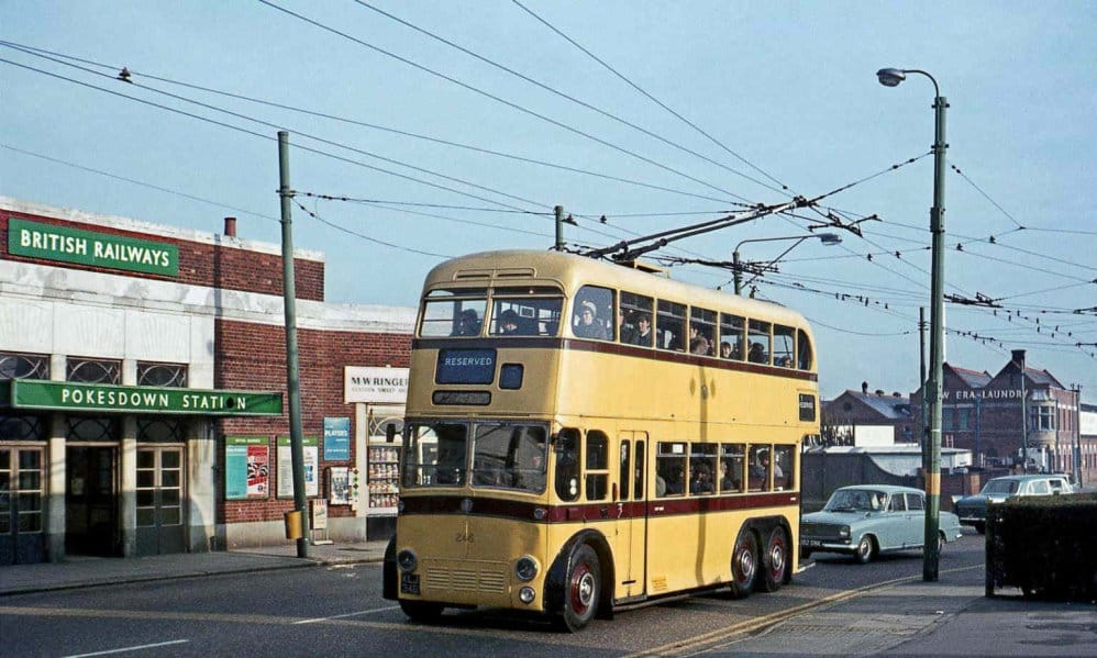 Colour photo of a yellow trolleybus driving past Pokesdown Station. The sign for Pokesdown Station is green. There is a blue car driving behind the bus. In the background a building with the sign New Era Laundry can be seen.
